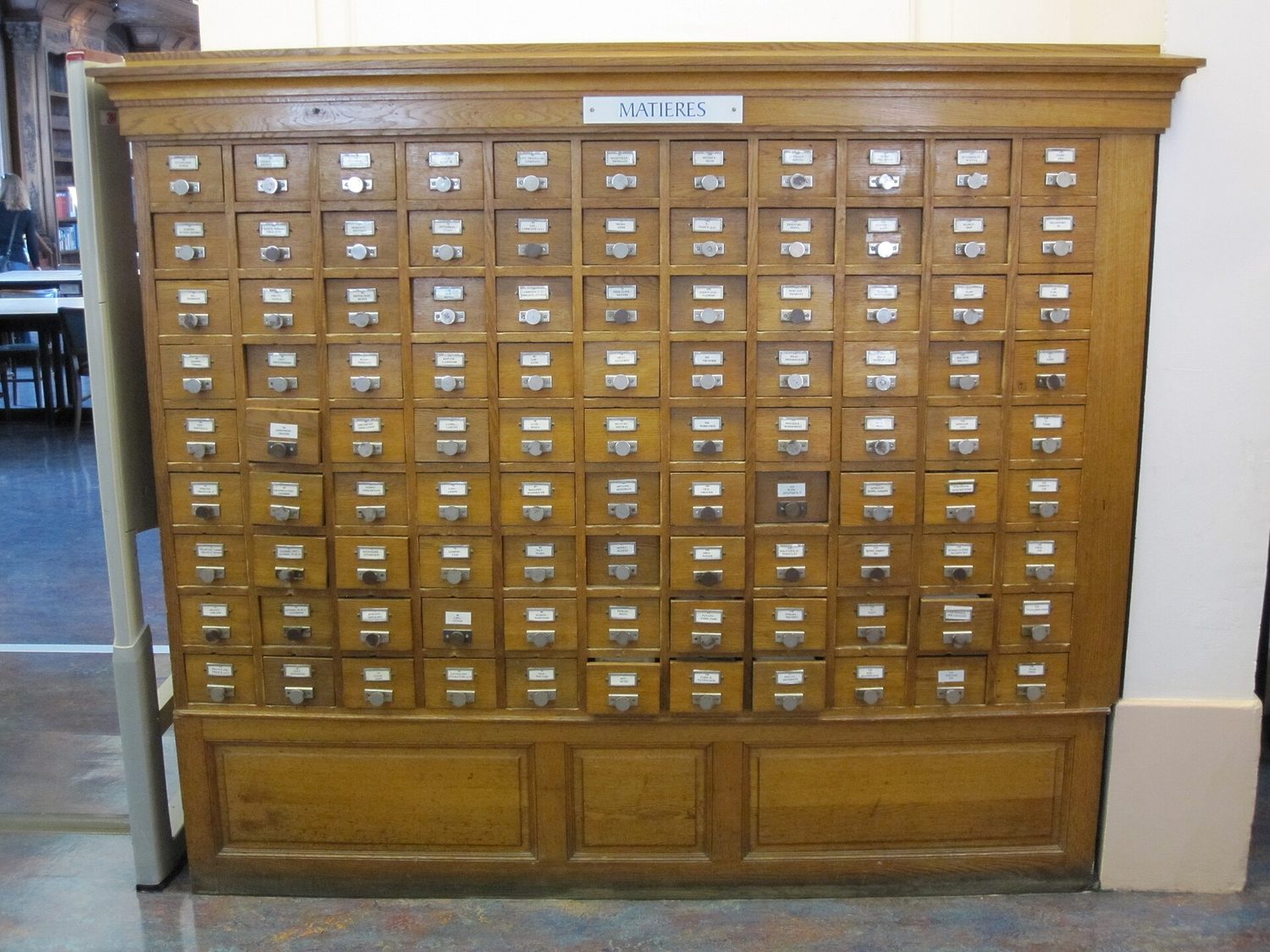 A library card catalogue: a wall of small wooden drawers, one pulled open to show the cards filed in order