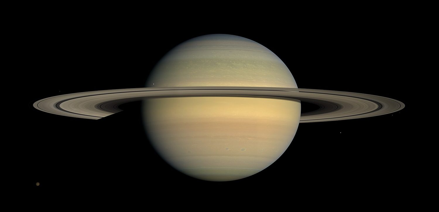 A NASA photograph of Saturn against black space: the round planet with pale cloud bands, surrounded by its wide flat ring system, with a few small moons visible as bright dots