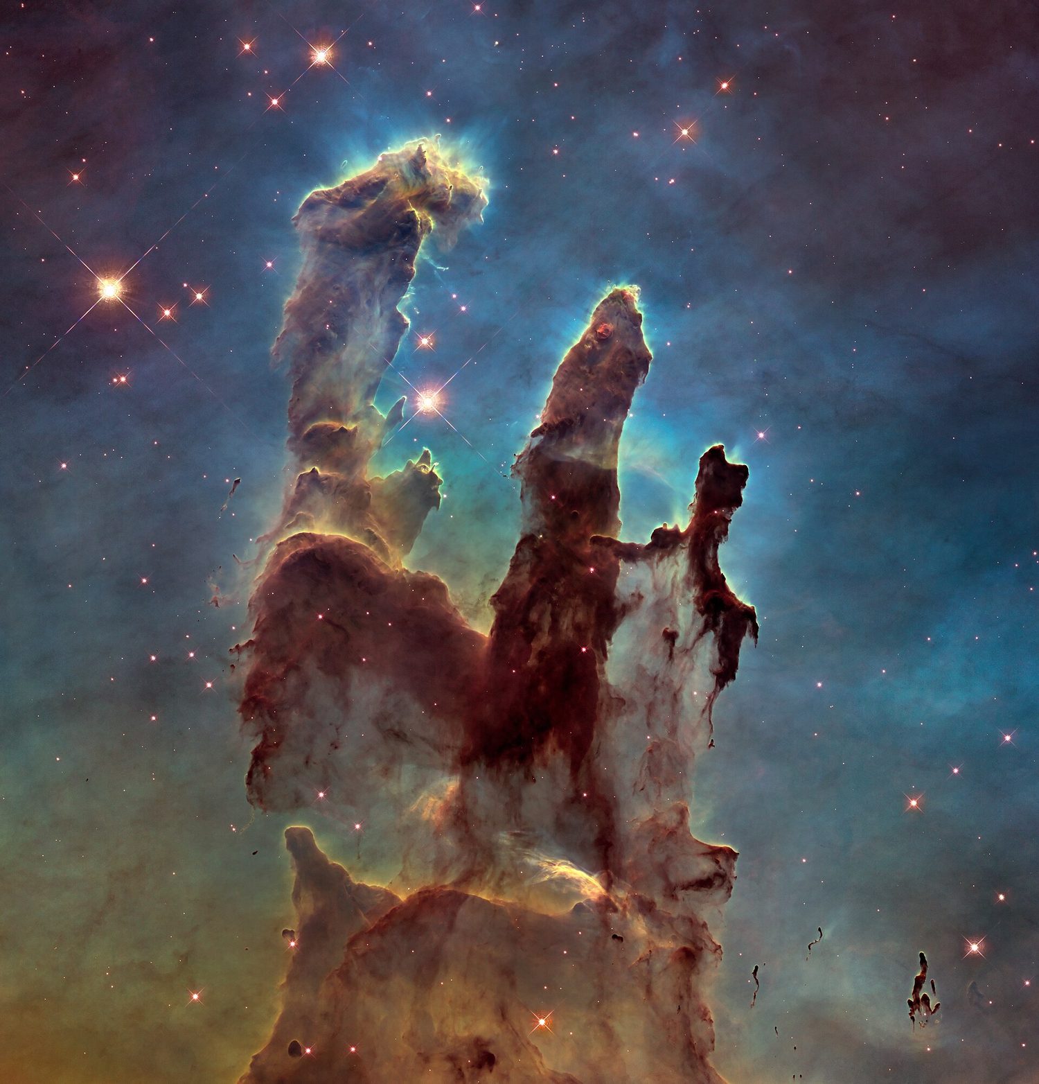 Three towering columns of brown and gold gas and dust rising against a blue-green nebula, tipped with bright young stars and scattered points of light